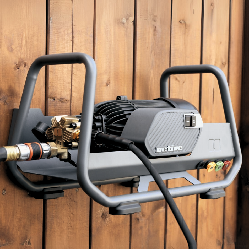 A Active™ 2.3 Electric Pressure Washer mounted against a wooden wall, featuring a black motor, metal frame, and brass fittings with a hose attached, delivering 2.3 GPM at 1100 PSI.